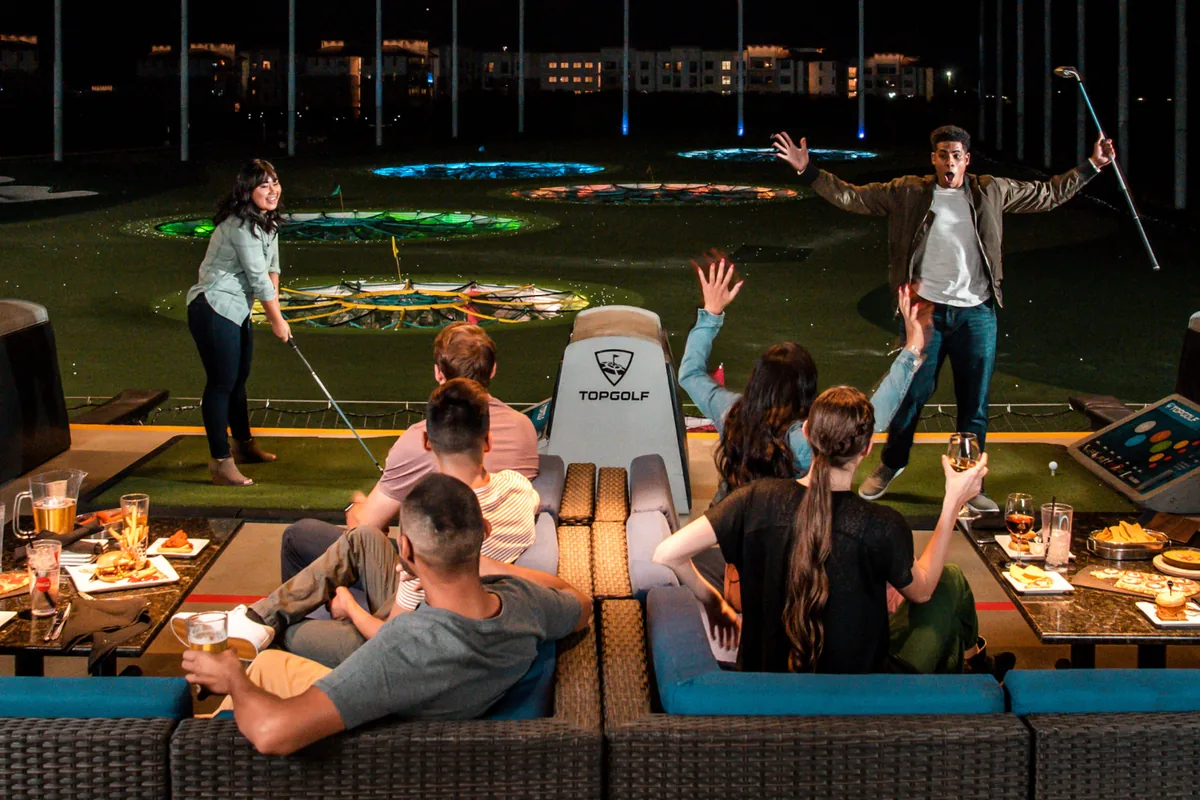 Gruppe in ihrer eigenen Bay bei Topgolf: Lounge-Sofas, Food & Drinks, zwei Spieler am Abschlag