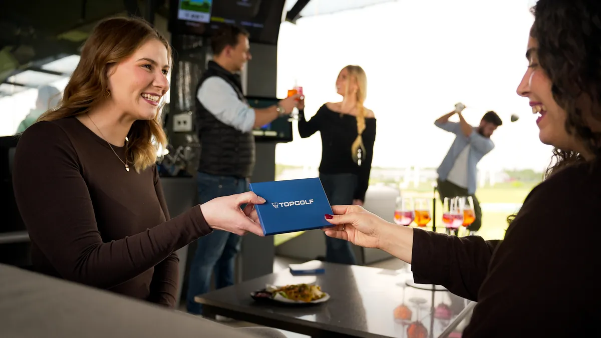 Ein Topgolf Geschenk wird in der Bay überreicht, Freunde stoßen im Hintergrund an